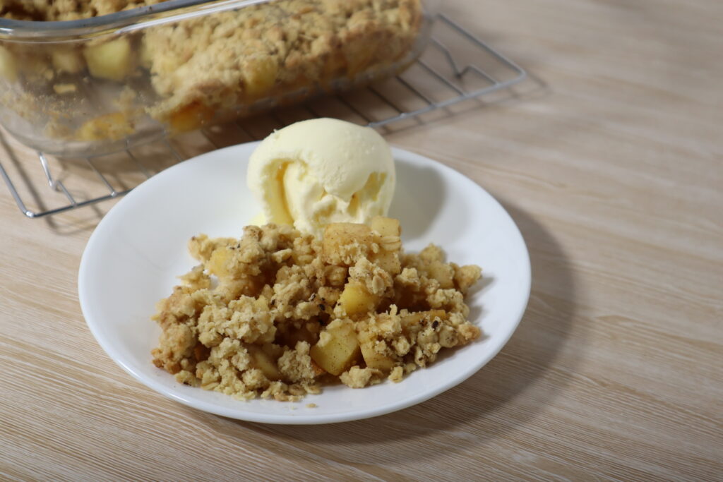 Side plate with a serve of apple crumble and a scoop of vanilla ice cream. background has a baking dish with cooked Apple Crumble.