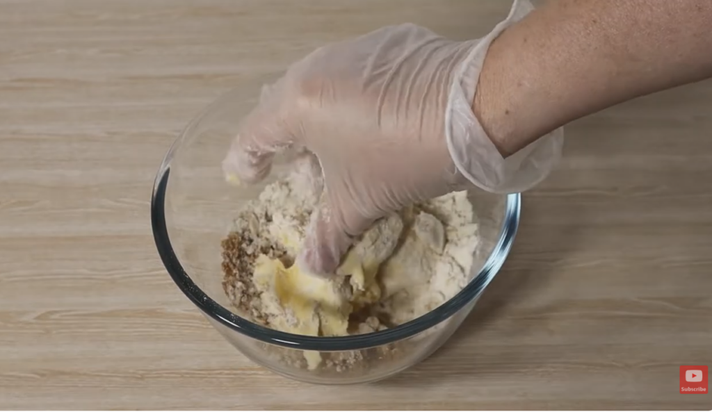 A glass bowl on a bench with crumble mixture for Apple Crumble, with a hand mixing it.