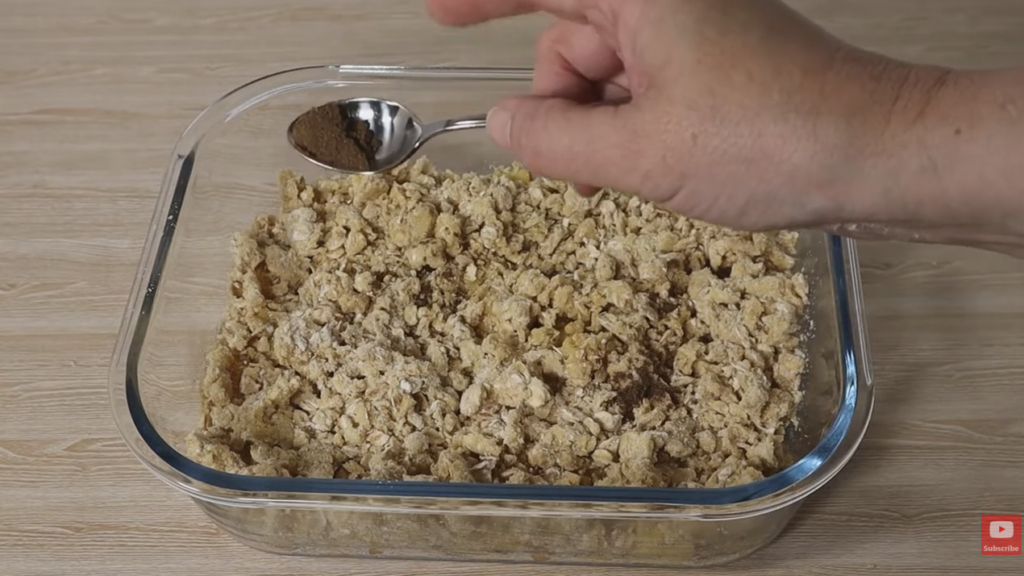 Glass baking dish with apples and crumble mixture with a hand sprinkling nutneg over the top.