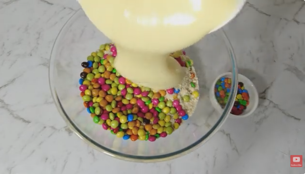 glass bowl with crushed biscults, M&M and pouring condensed milk over the top.