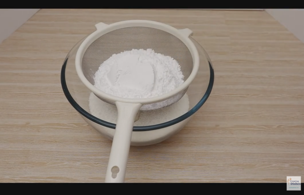 Large glass bowl with desiccated coconut and icing sugar in a sieve.