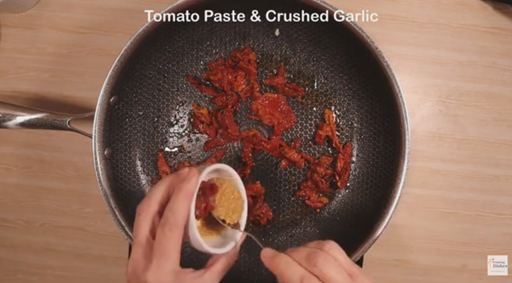 Frying pan with sundried tomato strips me adding tomato paste and crushed garlic to the pan.